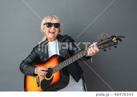 Senior woman in leather jacket and sunglasses studio standing isolated on gray playing guitar 48476343