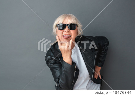 Senior woman in leather jacket and sunglasses studio standing isolated on gray showing tongue 48476344