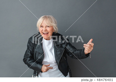 Senior woman in leather jacket studio standing isolated on gray playing imaginary guitar laughing 48476401