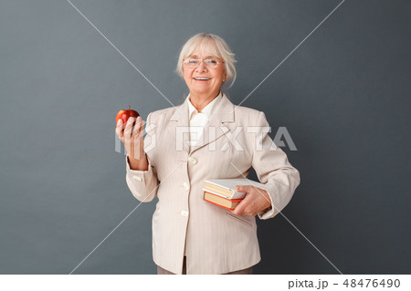 Senior woman in fromal suit and glasses studio standing isolated on gray with books and apple 48476490