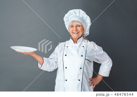 Senior woman chef studio standing isolated on gray holding plate looking camera laughing cheerful Senior woman chef studio standing isolated on gray holding plate looking camera laughing cheerful 48476544