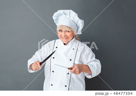 Senior woman chef studio standing isolated on gray with knives frightening playful Senior woman chef studio standing isolated on gray with knives frightening playful 48477304
