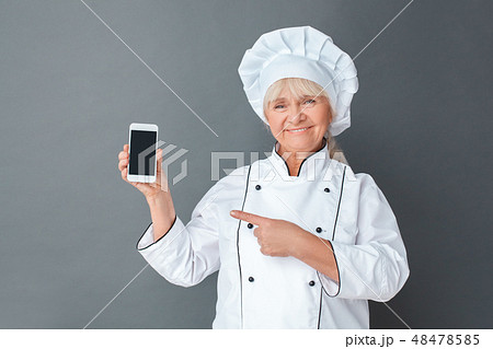 Senior woman chef studio standing isolated on gray with smartphone showing app on screen looking Senior woman chef studio standing isolated on gray with smartphone showing app on screen looking 48478585