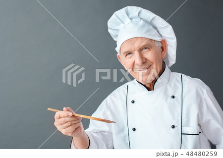 Senior chef studio standing isolated on gray holding spatula looking camera positive close-up Senior chef studio standing isolated on gray holding spatula looking camera positive close-up 48480259