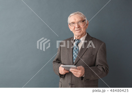 Senior man teacher wearing glasses studio standing isolated on gray holding digital tablet looking 48480425
