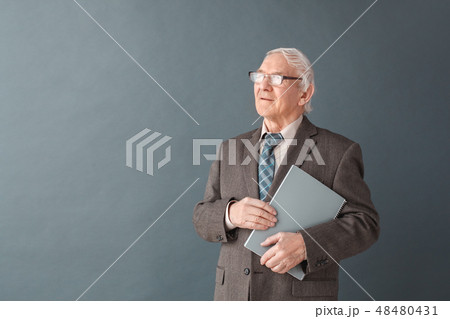 Senior man teacher wearing glasses studio standing isolated on gray holding book looking aside Senior man teacher wearing glasses studio standing isolated on gray holding book looking aside 48480431