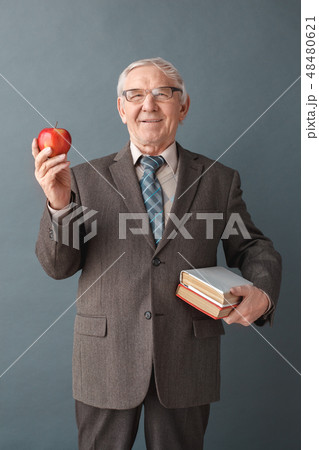 Senior man teacher wearing glasses studio standing isolated on gray with books and apple looking Senior man teacher wearing glasses studio standing isolated on gray with books and apple looking 48480621