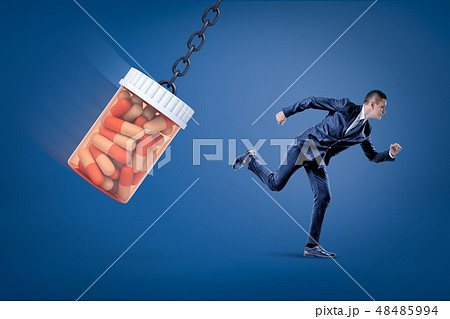 A side view of a businessman running from a huge full pills jar swinging on a chain on a blue A side view of a businessman running from a huge full pills jar swinging on a chain on a blue 48485994