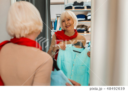 Emotional grey haired woman holding summer dress in front of store mirror 48492639