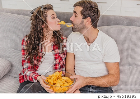 Smiling wife and man sitting on the couch. Watch the TV while eating chips in bed. Smiling wife and man sitting on the couch. Watch the TV while eating chips in bed. 48519395