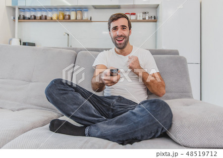 Beautiful hilarious young man holding a remote control. During this, the TV is watching while Beautiful hilarious young man holding a remote control. During this, the TV is watching while 48519712