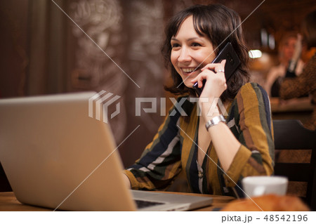 Pretty young woman smiling while talking on the phone and using her laptop in a coffee shop 48542196