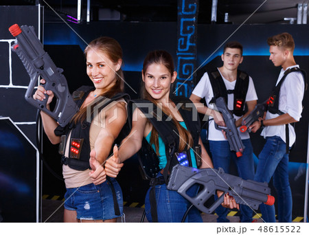 Two smiling girls posing with plastic laser pistols and two guys in background in laser tag room Two smiling girls posing with plastic laser pistols and two guys in background in laser tag room 48615522