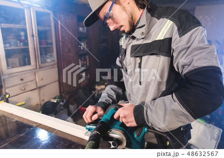 Young carpenter with a beard working with an electric plane with suction of sawdust. Leveling and 48632567