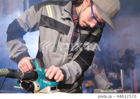 Young carpenter with a beard working with an electric plane with suction of sawdust. Leveling and 48632570