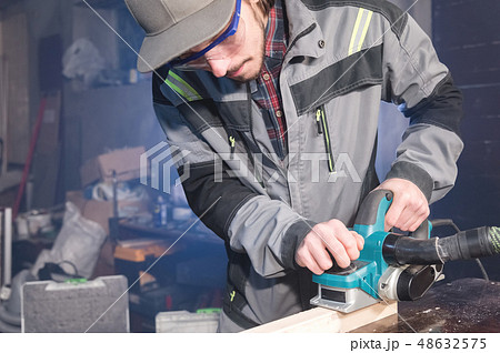 Young carpenter with a beard working with an electric plane with suction of sawdust. Leveling and Young carpenter with a beard working with an electric plane with suction of sawdust. Leveling and 48632575