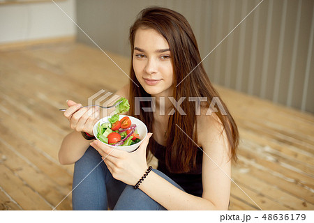 Sports beautiful girl sitting on the floor and eating a salad from fresh vegetables and greens Sports beautiful girl sitting on the floor and eating a salad from fresh vegetables and greens 48636179