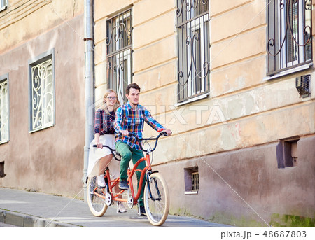 Young tourist couple, handsome man and pretty blond woman riding tandem bicycle along city street. 48687803