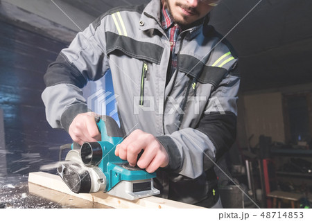 Young carpenter with a beard working with an electric plane with suction of sawdust. Leveling and 48714853