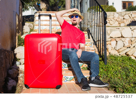 Holiday, travel and tourism concept - Handsome man with red suitcase sitting on a stairs 48725537