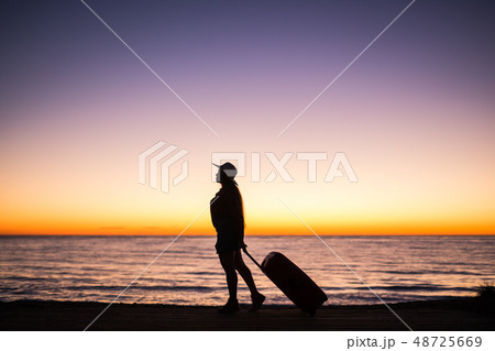 Traveling, beach and holiday concept - Female silhouette walking along ocean coast pulling suitcase Traveling, beach and holiday concept - Female silhouette walking along ocean coast pulling suitcase 48725669