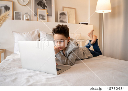 A little boy sitting on a big bed with a laptop book and looks into the camera. 48730484