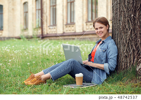Young student at university campus sitting under the tree browsing web on laptop looking camera 48742537