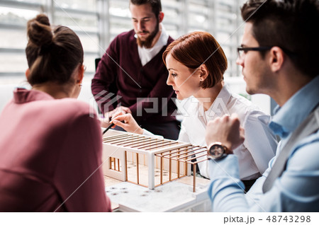 Group of young architects with model of a house standing in office, talking. Group of young architects with model of a house standing in office, talking. 48743298