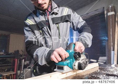 Young carpenter with a beard working with an electric plane with suction of sawdust. Leveling and Young carpenter with a beard working with an electric plane with suction of sawdust. Leveling and 48767406