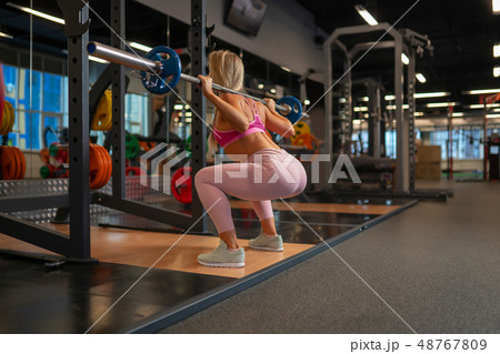 Photo from back of blonde girl crouching with barbell on her shoulders in gym. 48767809
