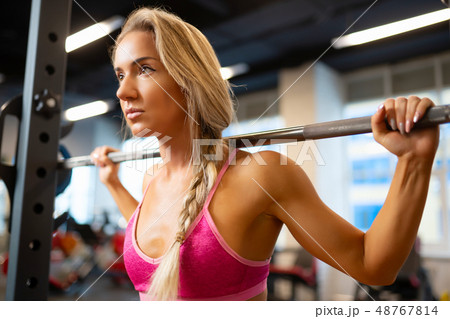 Portrait of sporty blonde standing with barbell on shoulders in gym. 48767814
