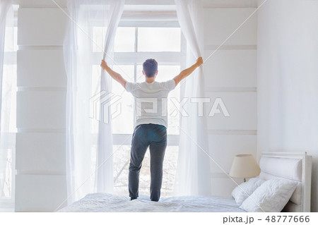 Young man opening window curtains in white bedroom in the morning 48777666