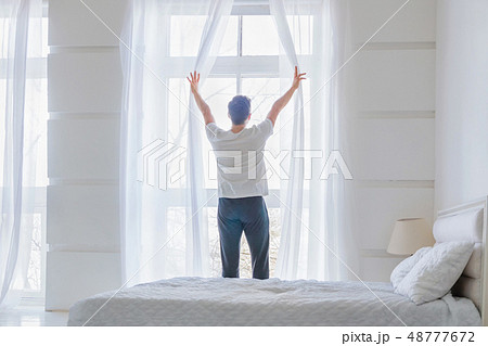 Young man near window at home. Man opening curtains in abstract white bedroom 48777672