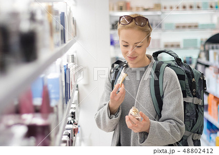 Blond young female traveler wearing travel backpack choosing perfume in airport duty free store. Blond young female traveler wearing travel backpack choosing perfume in airport duty free store. 48802412