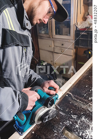 Young carpenter with a beard working with an electric plane without suction of sawdust. Leveling and Young carpenter with a beard working with an electric plane without suction of sawdust. Leveling and 48832207