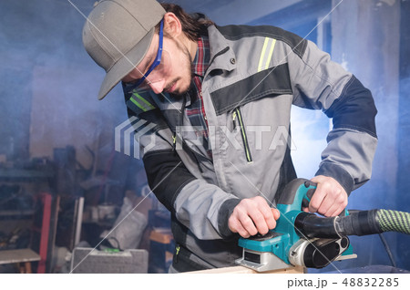 Young carpenter with a beard working with an electric plane with suction of sawdust. Leveling and 48832285