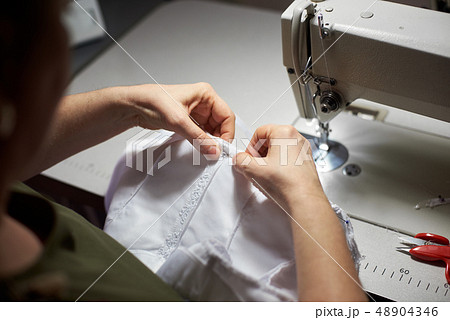 Needlewoman draping white garment with needle and her hands. Sewing machine, scissors on background 48904346