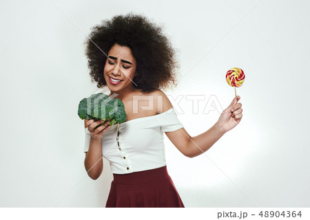 I choose healthy food! Pretty afro american woman is facing a choice of healthy broccoli or lollipop 48904364