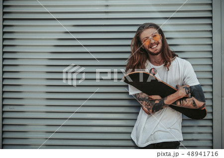 Handsome hipster guy with long hair holding skateboard Handsome hipster guy with long hair holding skateboard 48941716