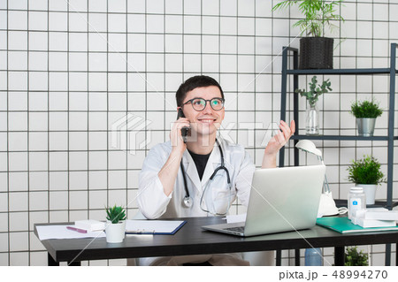 Male doctor using telephone while working on computer at table in clinic Male doctor using telephone while working on computer at table in clinic 48994270