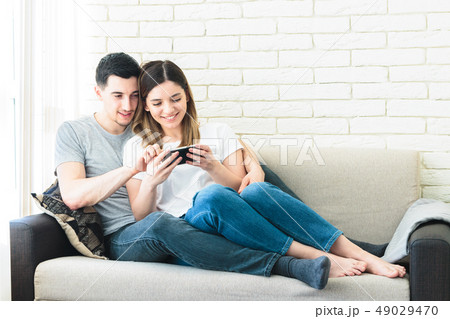 Relaxed couple using a generic mobile phone together sitting on a sofa in the living room at home 49029470
