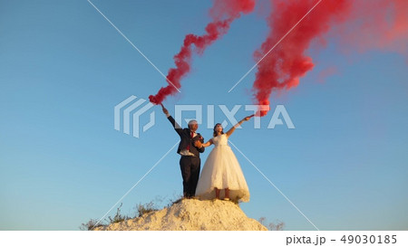 Happy bride and groom waving colored pink smoke against blue sky and laughing. Honeymoon. Romance 49030185