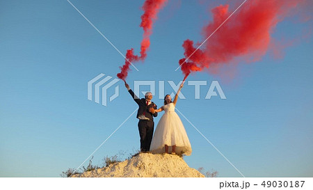 Happy bride and groom waving colored pink smoke against blue sky and laughing. Honeymoon. Romance 49030187