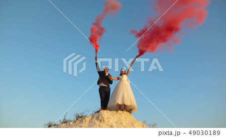 Happy bride and groom waving colored pink smoke against blue sky and laughing. Honeymoon. Romance 49030189