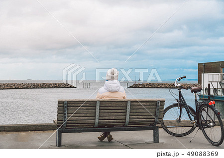 A young Caucasian woman sits with her back on a wooden bench overlooking the Baltic Sea on the 49088369