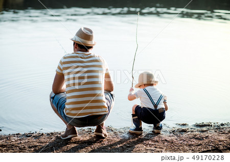 A rear view of mature father with a small toddler son outdoors fishing by a lake. A rear view of mature father with a small toddler son outdoors fishing by a lake. 49170228