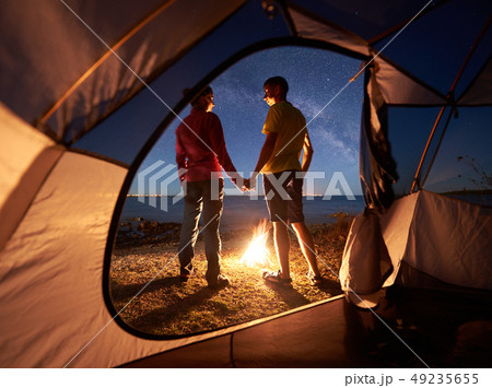 Young couple man and woman having rest at tourist tent and burning campfire on sea shore near forest 49235655