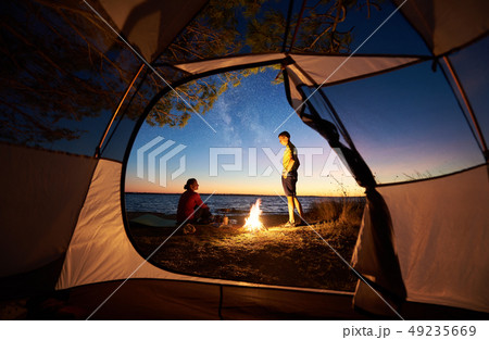 Young couple man and woman having rest at tourist tent and burning campfire on sea shore near forest Young couple man and woman having rest at tourist tent and burning campfire on sea shore near forest 49235669