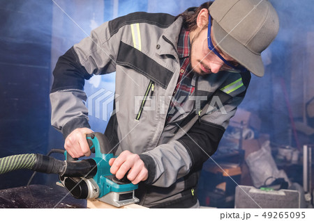 Young carpenter with a beard working with an electric plane with suction of sawdust. Leveling and 49265095