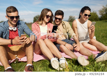 smiling friends with smartphones sitting on grass 49284284